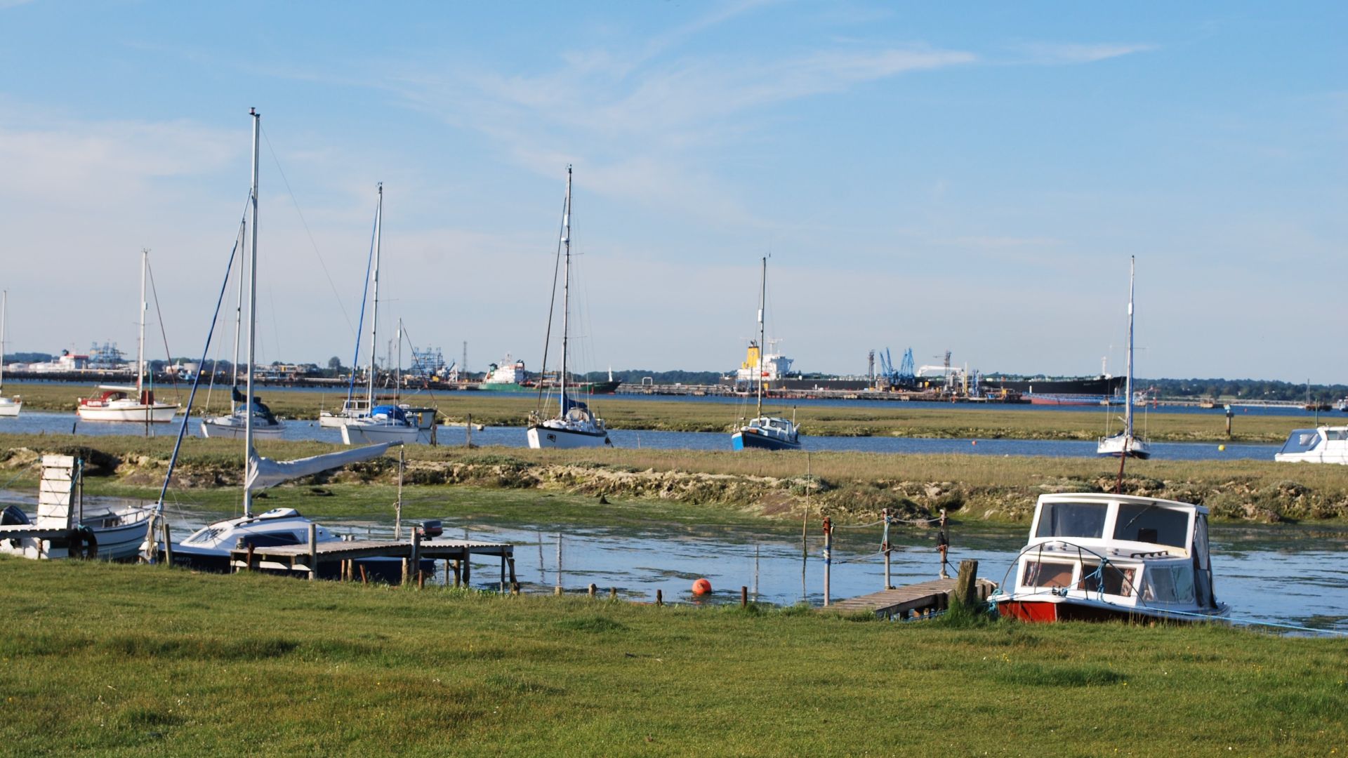 Boats moored on Ashlett Creek at low tide, with grassy marsh and distant industrial port under a blue sky
