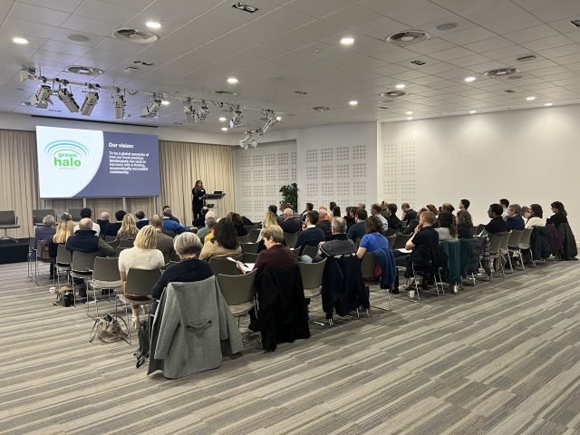 Audience listening to a speaker beside a screen showing the Green Halo logo at the first Green Halo Conference