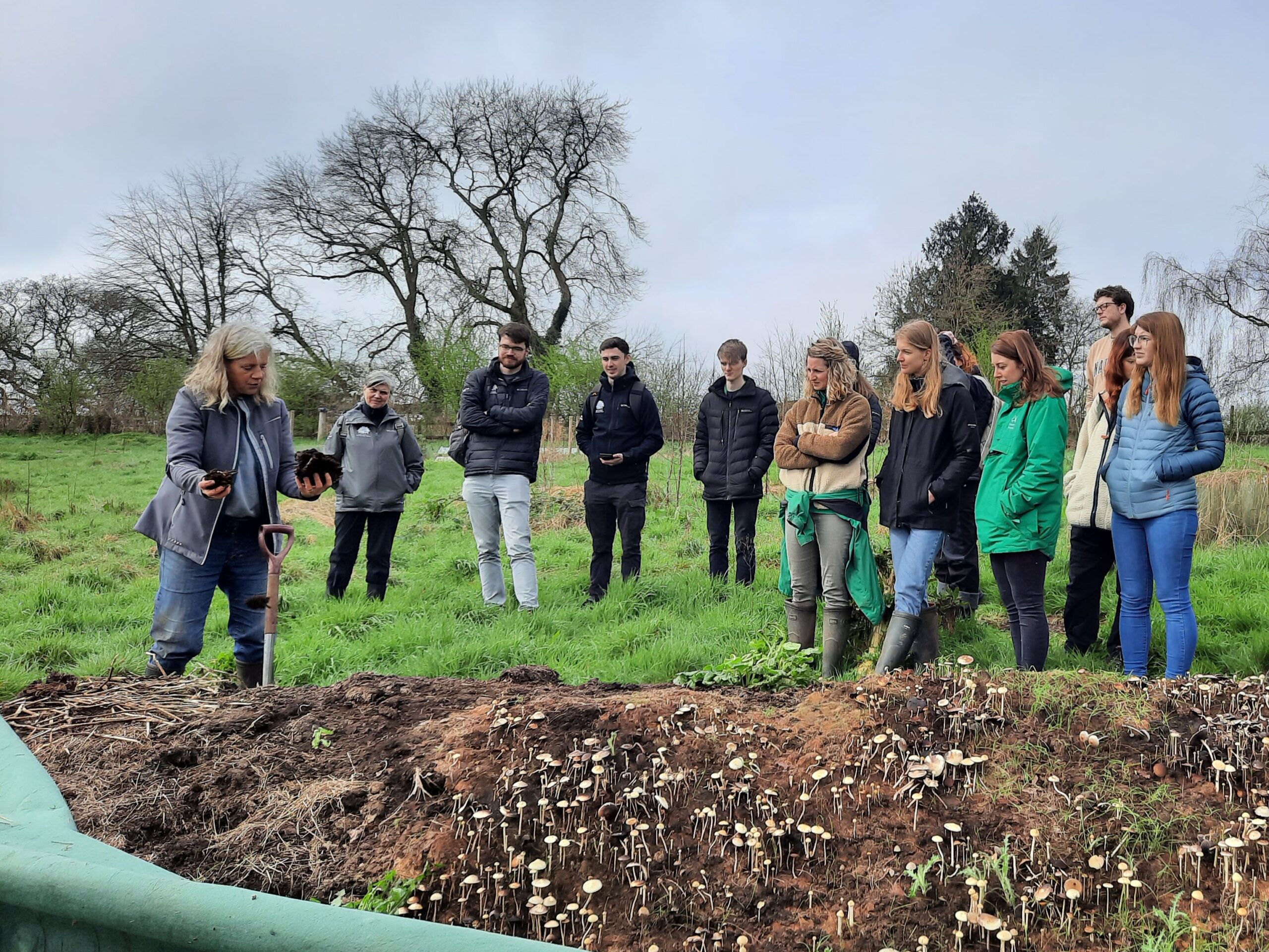 Farm instructor shows soil samples to a group in a field beside a compost pile covered with small mushrooms