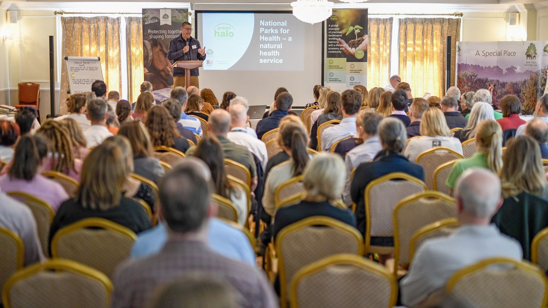 Speaker at podium addresses seated audience at Green Halo Conference 2024, with slide reading National Parks for Health on screen