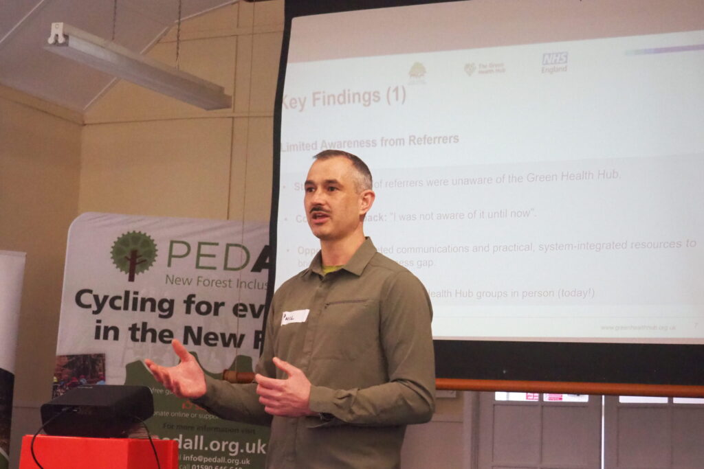 Health fellow Phil Godfrey speaking at a presentation, gesturing in front of a projected slide and a Pedall cycling banner