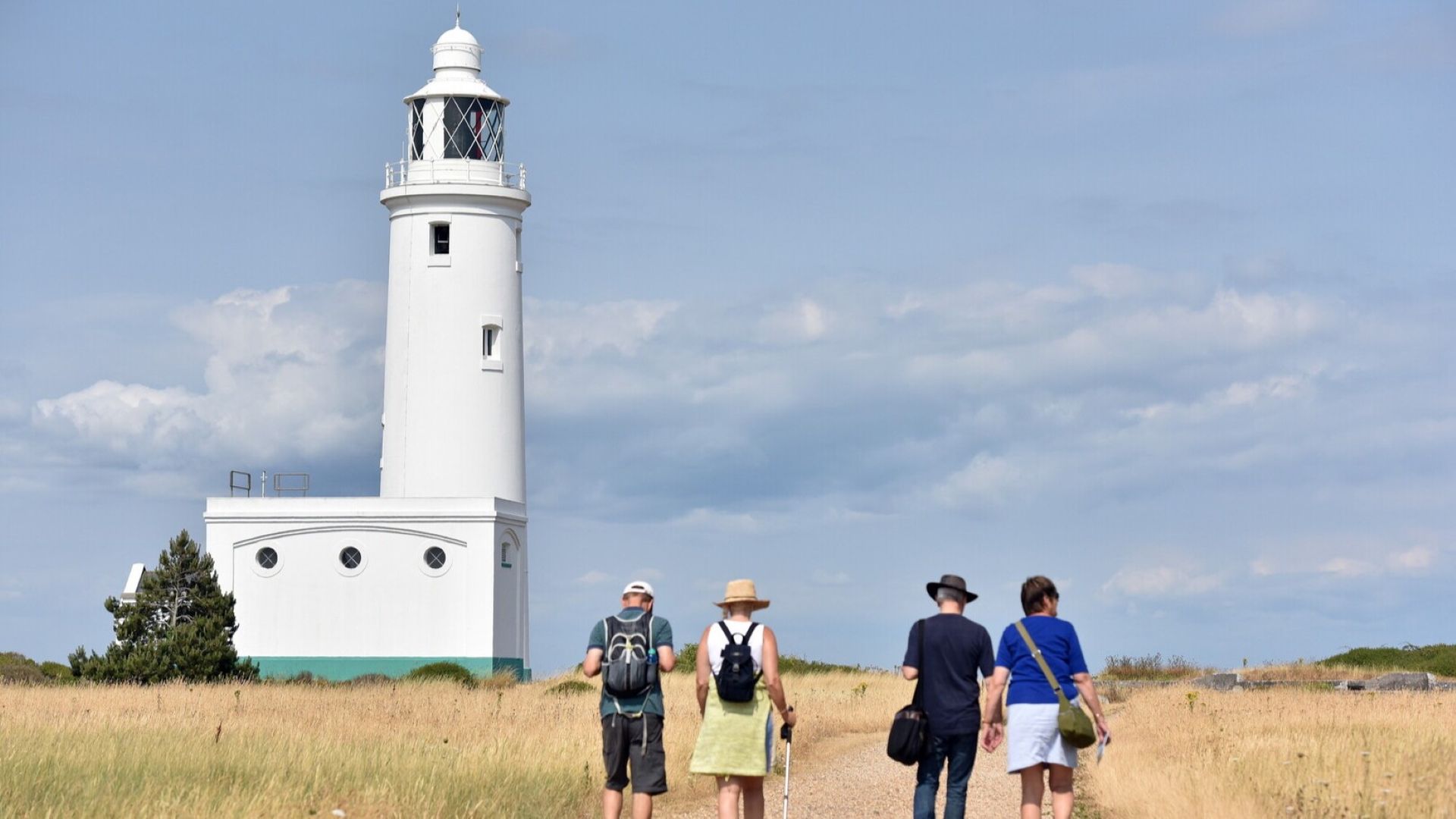 Hurst Point Lighthouse, a white tower by a grassy path, with four visitors walking toward it under a cloudy sky