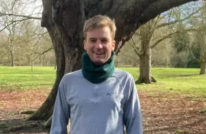 Smiling man wearing a green scarf and grey top standing in a park beside a large tree