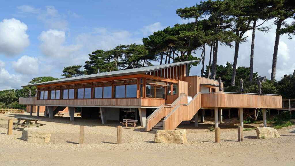 Lepe Beach visitor centre and cafe, a modern timber building on stilts with large windows, stairs and terrace among pine trees
