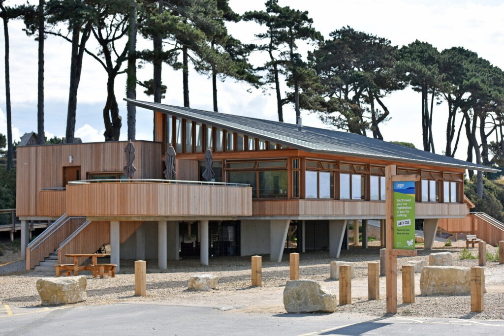 Modern wooden Lepe Beach cafe on stilts with slanted roof, terrace, and pine trees behind, viewed from the car park
