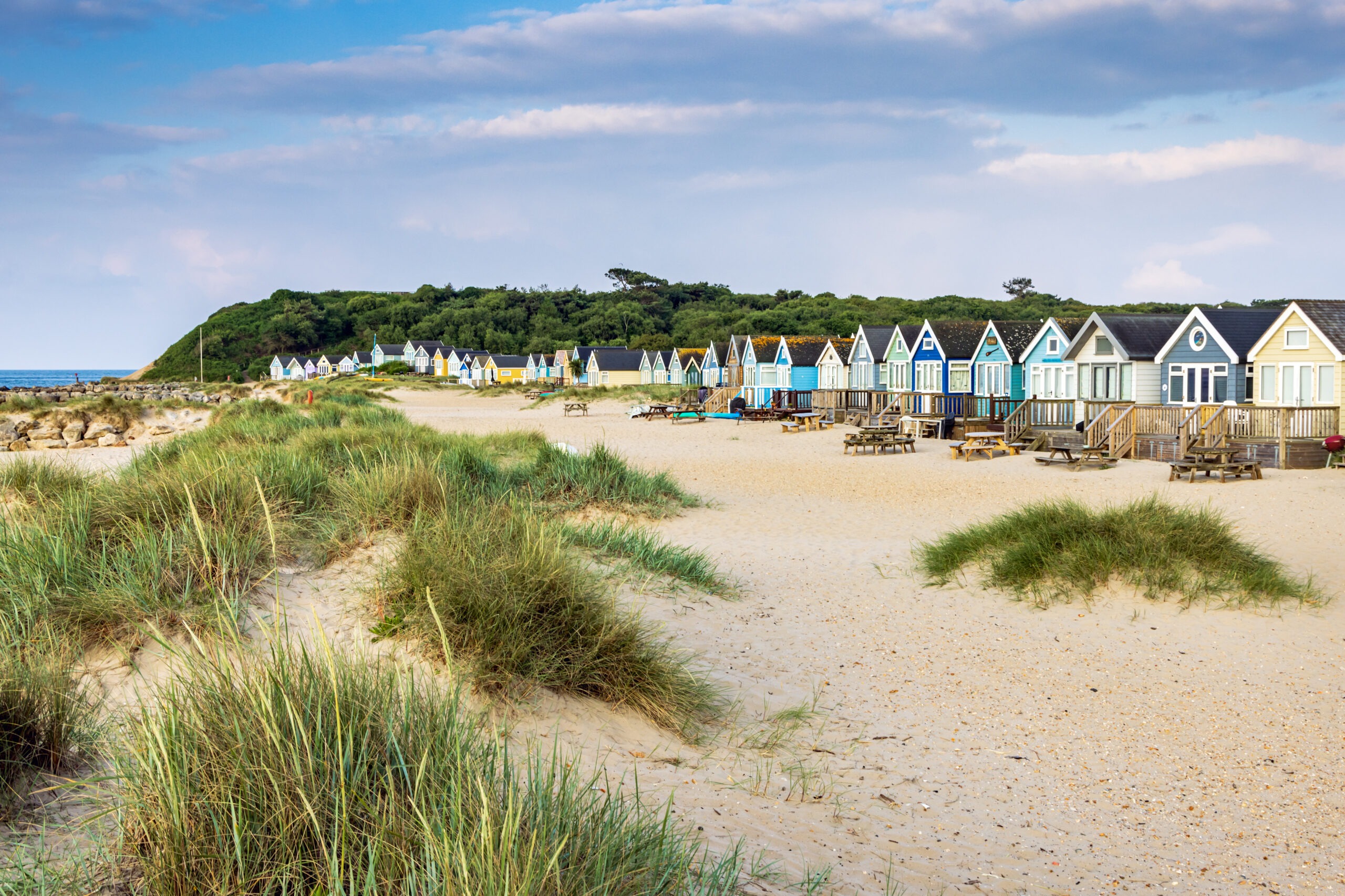Colourful beach huts on Mudeford Spit, Dorset, lining a sandy beach with dunes, picnic benches and a cloudy sky