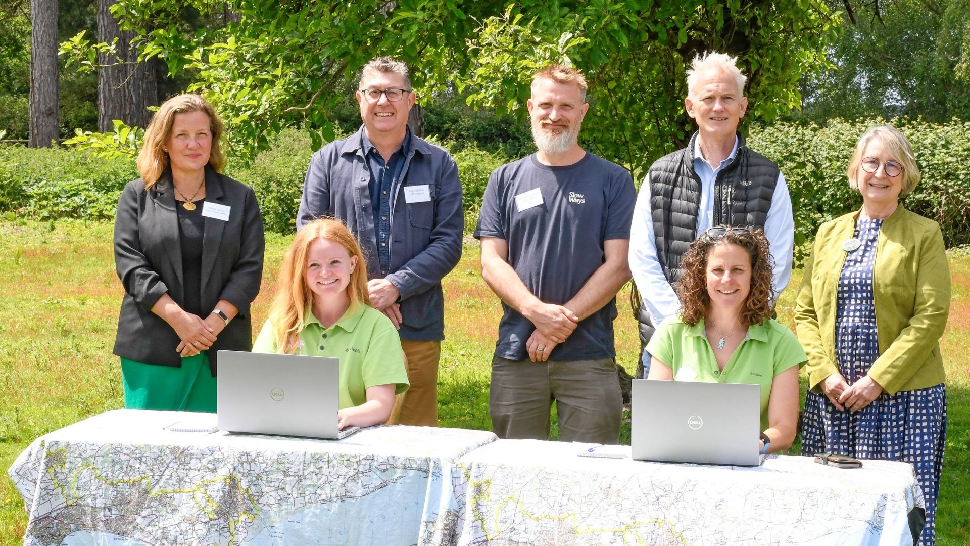 Six people at Outdoor Office Day UK launch, two seated at laptops on a map-covered table in a sunny park with trees behind