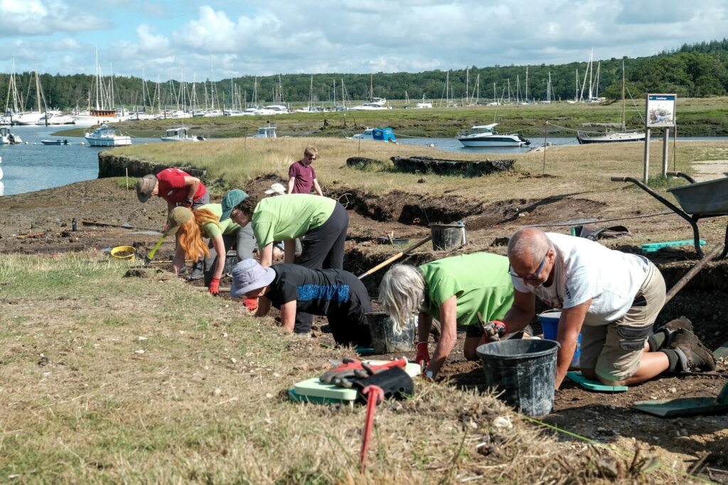 Volunteers kneel and dig in an excavation trench beside a harbor, with sailboats and a marina in the background