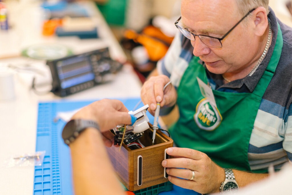 Man in green apron repairs electronics in a wooden box with a probe while another person holds components at a workbench