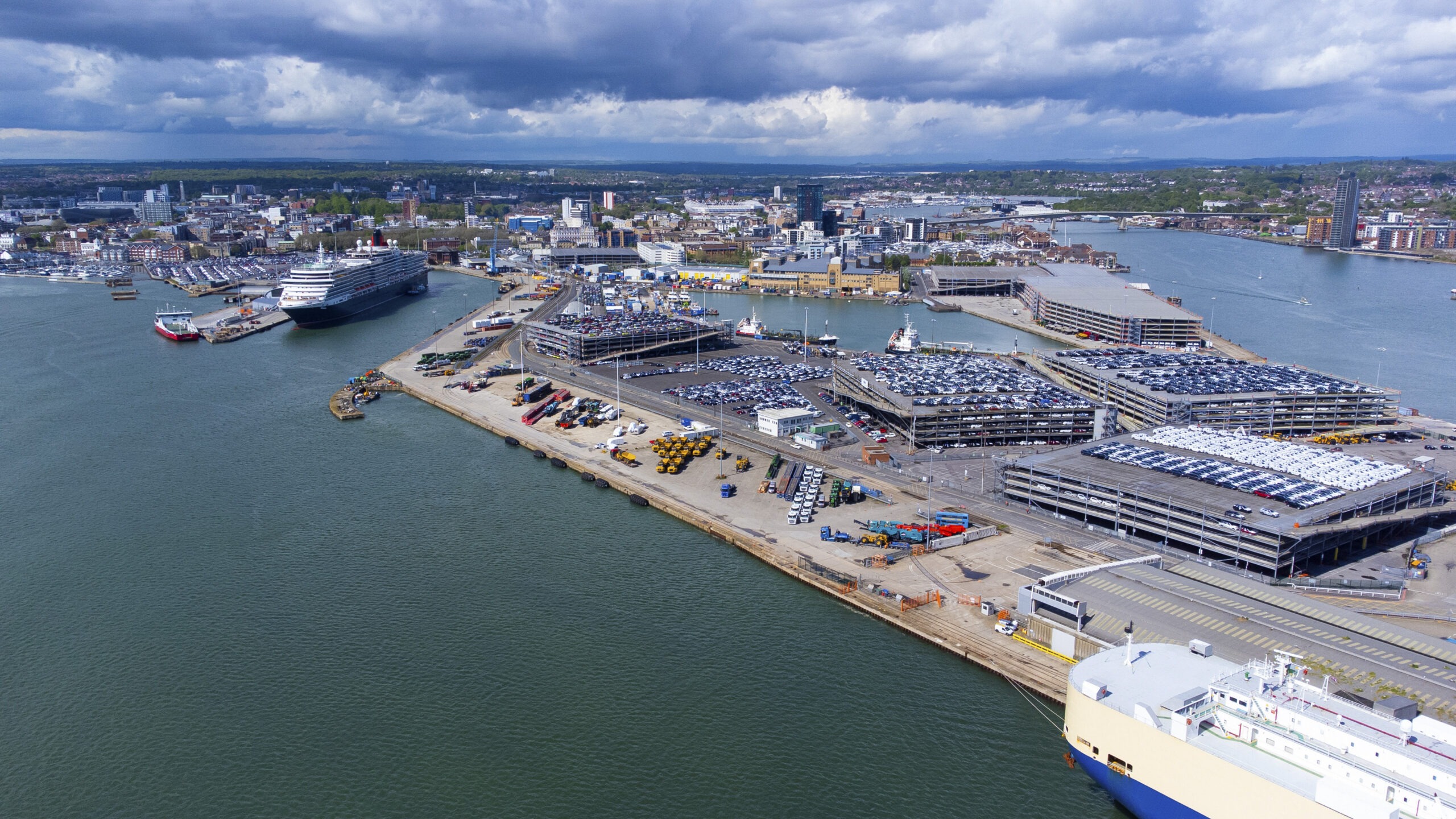Aerial view of the Port of Southampton docks with a cruise ship, car storage lots, and waterfront buildings under cloudy sky