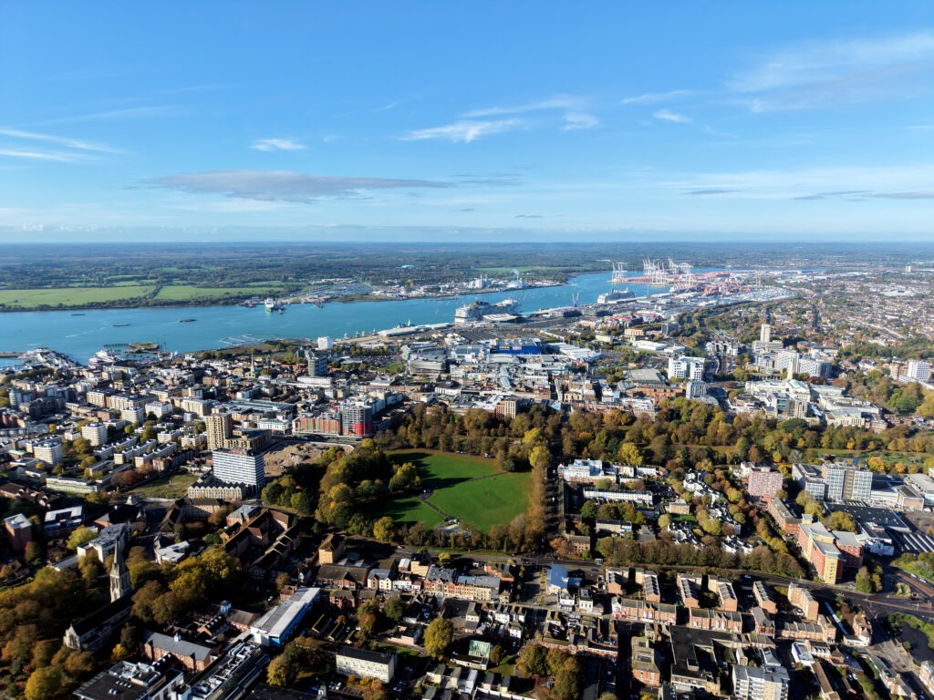 Aerial view of Southampton, England, showing city centre buildings, a riverside port with cranes, and cruise ships on the waterway