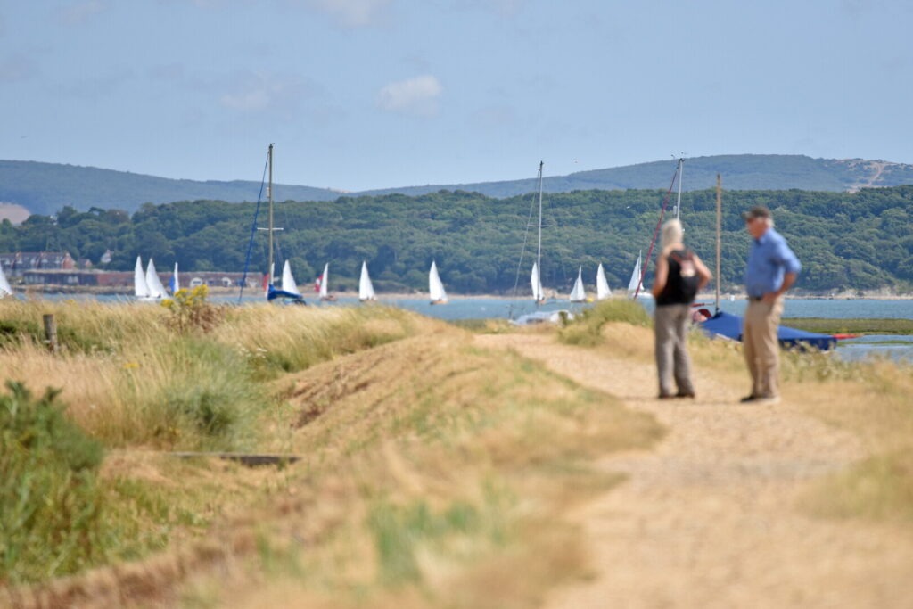Two walkers on a coastal path near Keyhaven Marshes, with sailboats on the water and green hills in the background